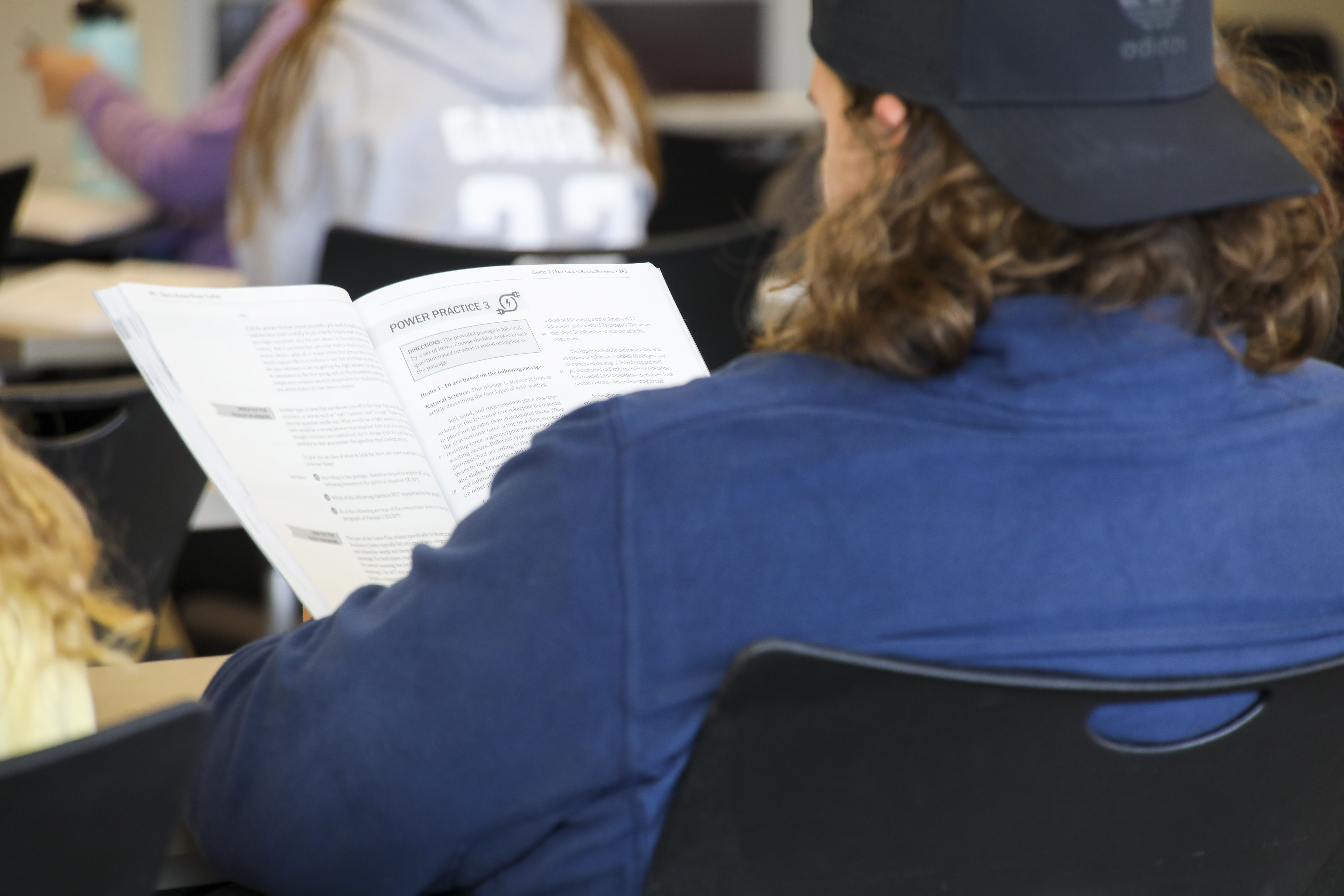 The back of a student who is in a classroom reading a workbook